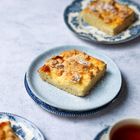 Ein Stück Streuselkuchen liegt auf einem blauen Teller. Im Hintergrund sind weitere Kaffeetassen und Teller zu sehen.