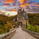 Eine gepflasterte Brücke führt zu einer mittelalterlichen Burg, umgeben von einem bewaldeten Hang. Der Himmel ist orange-violett.