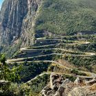Serpentinenstraße schlängelt sich steil entlang eines grünen, felsigen Berghangs. Unten dichte Vegetation. Gestaffelte Kurven.