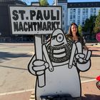 "ST. PAULI NACHTMARKT" auf einem Schild, gehalten von einer gemalten Cartoonfigur. Eine Frau steht lächelnd daneben.