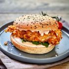 Ein Burger mit Sesamnatur und Schwarzkümmel auf dem Brötchen, knuspriges Hähnchen, Salat und Soße auf einem grauen Teller.
