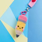 Ein gehäkelter Bleistiftanhänger mit Gesicht und herzförmigem Clip vor buntem Hintergrund.