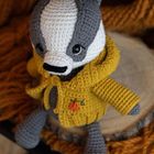 Ein gehäkelter Dachs in grauen und weißen Tönen trägt eine gelbe Strickjacke mit einer kleinen Kürbis-Stickerei.