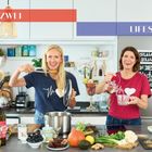 "TEIL ZWEI", "LIFESTYLE" auf farbigen Balken. Zwei fröhliche Frauen kochen in moderner Küche, umgeben von frischen Zutaten.