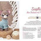 "Sophie, die Katze mit Pullower." Eine gehäkelte Katze mit blauem Pulli sitzt auf einem Rattanuntergrund, umgeben von Strohkugeln.