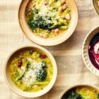 Keine Texte erkennbar. Drei Schalen mit gelber Suppe, Gemüse und geriebenem Käse auf einem Tuch. Eine Schale mit lila Mischung.