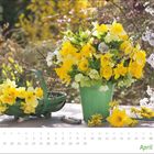 April. Ein grüner Topf mit gelben Blumen auf einem Tisch. Links ein Korb mit ähnlichen Blumen. Blühende Zweige im Hintergrund.