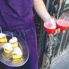 Zwei Gläser mit rotem Getränk, eine Person in lila Schürze hält ein Tablett mit kleinen Häppchen. Im Hintergrund ist eine alte Holztür.