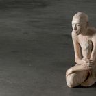 Rechts beugt sich eine menschenähnliche Skulptur aus hellem Material nach vorne, in hockender Position.