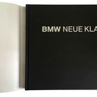 Text: "BMW NEUE KLASSE". Ein geöffnetes Buch mit schwarzem Umschlag und weißer Schrift. Links ist eine leere Seite sichtbar.