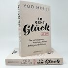 Zu sehen sind die Bücher "So geht Glück" von Yoo Min Ji, aufrecht stehend und liegend.