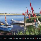 4 April. Zwei Boote am Seeufer. Rote Fahnen wehen neben einem Steg, umgeben von Schilf und ruhigem Wasser.