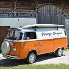 Text: "Vintage Camper". Ein orange-weißer Oldtimer-Camper steht vor einem Holztor. Surfbretter auf dem Dach.