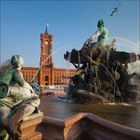 Ein rotes Backsteingebäude mit Uhrturm, davor ein Brunnen mit mythologischen Figuren und sprudelndem Wasser.