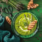 Eine grüne Spargelsuppe in einer Schale, garniert mit Sahne, Spargel und geröstetem Brot auf einem Holzuntergrund.