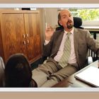 Ein Mann im Anzug sitzt lässig im Büro, Füße auf dem Tisch, Zigarettenstummel in der Hand, hinter ihm ein Schrank.