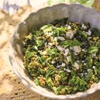 Ein weißer Teller mit grünem Salat, Feta und Kernen, auf einer Spitzen-Tischdecke, mit drei grünen Gläsern im Hintergrund.