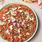 Ein Teller mit Tomaten-Linsengericht, garniert mit Feta, Granatapfelkernen und Pistazien. Daneben sind Gläser und Fladenbrot.