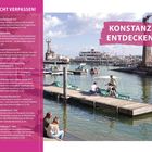 "Nicht verpassen!", "Konstanz entdecken". Hafen mit Booten, Promenade, Menschen, im Hintergrund Statuen und ein großes Schiff.