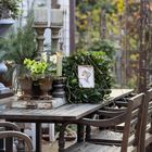 Tisch mit grünen Pflanzen und Kerzen. Ein kleiner gerahmter Engel ist in der Mitte platziert. Rustikale Gartenstühle.