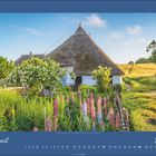 April 2027. Ein strohgedecktes Haus mit blühendem Garten, blauer Himmel und sanfte Hügel im Hintergrund.