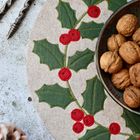 Stickerei mit Stechpalmenblättern und roten Beeren auf einem runden Stoff. Daneben Nüsse in einer Schale.