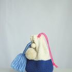 Ein gestricktes Täschchen in Weiß und Blau mit rosa Kordel, Pompons und einer blauen Quaste auf einem hellen Hintergrund.