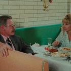 Zwei Personen sitzen in einem Restaurant an einem Tisch mit leergegessenen Tellern und Getränken in einem 70er-Jahre-Stil.