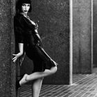 Ich kann den Text nicht erkennen. Eine Frau lehnt sich in einem schwarzen Kleid an eine Wand, mit glatten Haaren und High Heels.