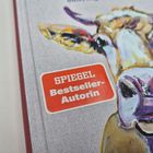Text: "SPIEGEL Bestseller-Autorin." Darstellung einer farbenfrohen Kuh in Nahaufnahme auf einem Buchcover.