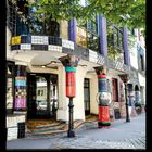 IV 2027. Unter einem grünen Blätterdach ist ein bunter, verspielter Gebäudeeingang mit unregelmäßigen Formen.