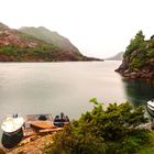 Keine Texte im Bild. Zwei Boote an einem kleinen Steg in einem ruhigen Fjord, umgeben von grünen Hügeln und Felsen.