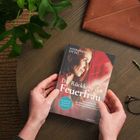 "Die Rückkehr der Feuerfrau" von Sandra Saviera Otto. Zwei Hände halten ein Buch auf einem Tisch mit Pflanze und Brille.