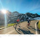 Text: "April", Kalenderdaten unten. Zwei Radfahrer auf Bergstraße, teilweise schneebedeckte Berge, Sonnenschein, Logo rechts.
