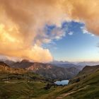 Ein kleines, blaues Bergsee liegt in grünen Hügeln, umgeben von majestätischen Bergen und dramatisch beleuchteten Wolken.