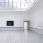 Zwei Gemälde an weißen Wänden in einem hellen Raum mit Dachfenster, Holzboden, und einer zentralen Türöffnung.