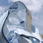 Eine große, blaue Skulptur mit geschwungenen, ineinander greifenden Formen vor einem bewölkten Himmel.