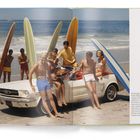 Eine Gruppe junger Menschen in Badekleidung posiert mit Surfbrettern auf einem weißen Cabrio am Strand.