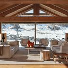 Ein gemütliches Wohnzimmer mit Holzbalken und großen Fenstern zeigt eine verschneite Berglandschaft im Hintergrund.