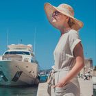 Seite 9. Eine Frau in Sommerkleidung und Hut steht neben einer großen Yacht im Hafen unter klarem blauem Himmel.