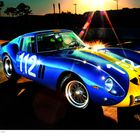 Zahl "112" auf blau-gelbem Sportwagen, Sonnenlicht reflektiert. Retro-Design, Bäume und Sonnenuntergang im Hintergrund.