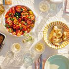 Ein Tisch mit gedeckten Tellern, Gläsern, Brot, Tomatensalat, gebratenem Fleisch und einer Flasche Öl in sonnigem Licht.