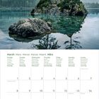 Kalender: März. Montag bis Sonntag auf sechs Sprachen. Oben ein ruhiger See mit Bäumen auf Felsen, neblige Berge im Hintergrund.