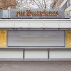 Zur Bratpfanne, Menütafeln links und rechts. Geschlossener Imbissstand mit Rollladen. Herbstliche Szene mit Bäumen.