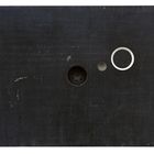 Dunkle, rechteckige Fläche mit drei runden Objekten: ein schwarzer Kreis, ein kleiner grauer Kreis und ein weißer Ring.