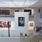 Großer Galerieraum mit moderner Kunst an weißen Wänden, mehrere farbige Gemälde, Treppe, Glasgeländer.