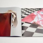 Ein offenes Buch zeigt links ein rotes Bild mit einem beigen Stoff, rechts eine Hand unter rosa Papier auf schwarz-weißem Boden.