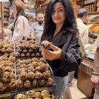 Eine Frau in einem Laden hält eine Süßigkeit mit Nüssen. Hinter ihr sind Verkäufer und bunte Gewürze.