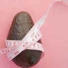 Zahl "015" unten. Eine Avocado ist von einem Maßband umwickelt, das die Zahlen 53 bis 112 zeigt, auf rosa Hintergrund.