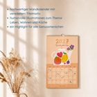 hochwertiger Wandkalender, humorvolle Illustrationen, Kalenderblätter, Vase mit Trockenblumen, Lichteinfall auf Wand.